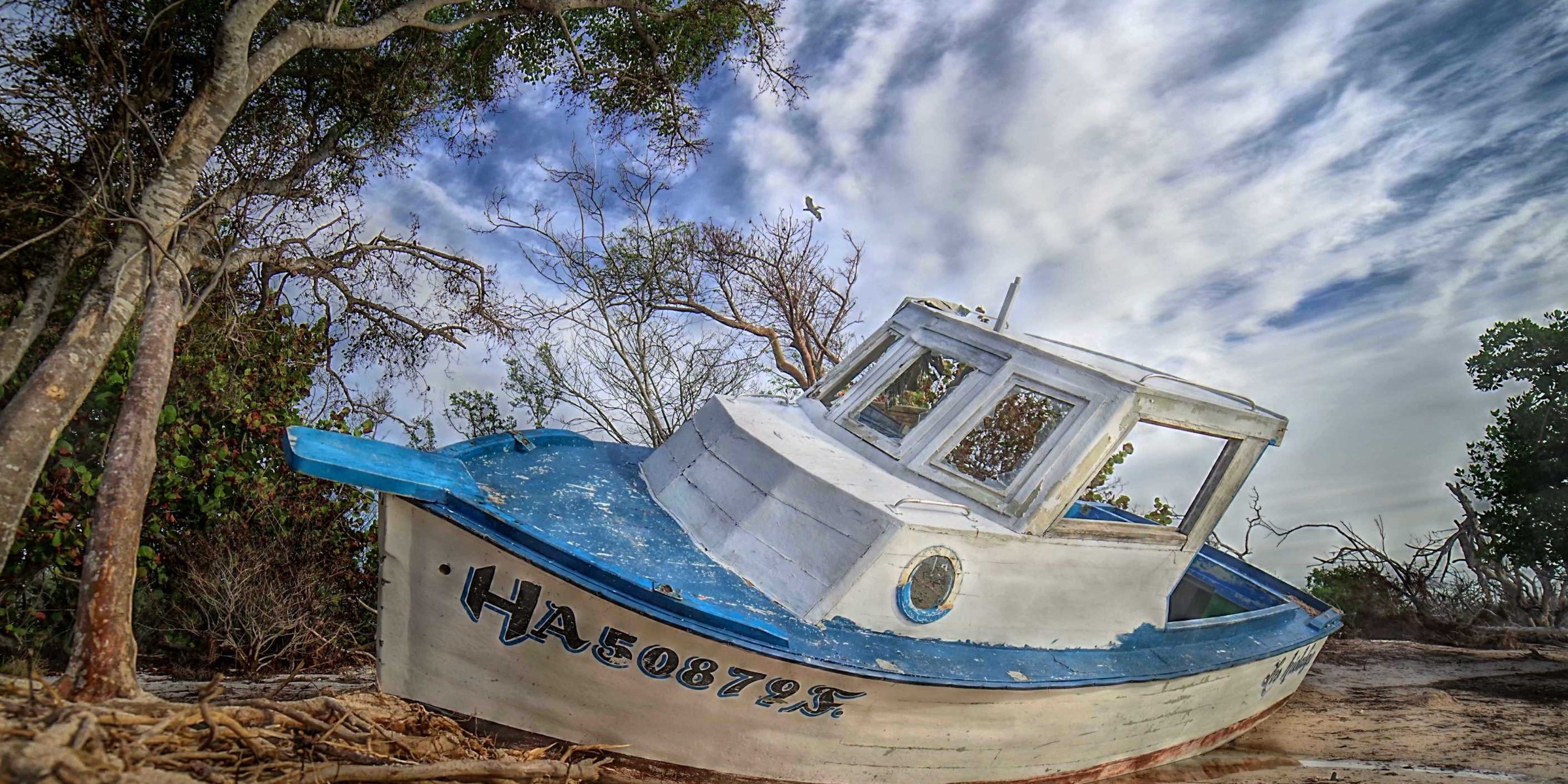 A weathered blue-and-white fishing boat grounded on a sandy beach under an expressive cloud-filled sky.