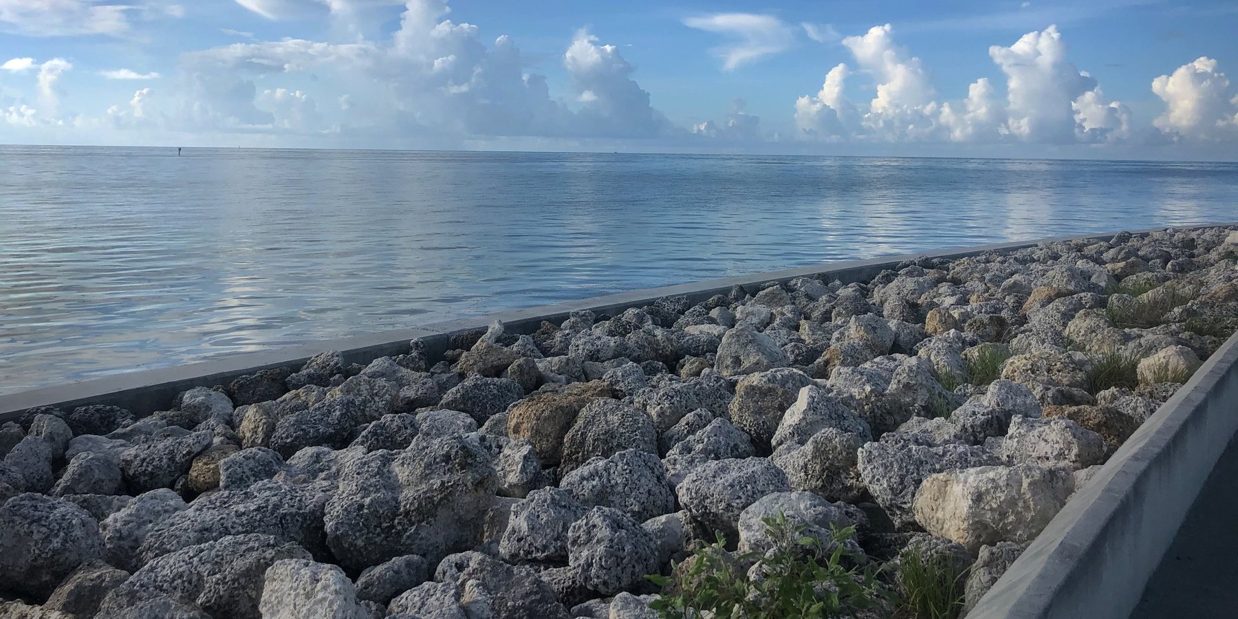 Photograph of a calm sea under tall clouds, seen across a rocky breakwater.