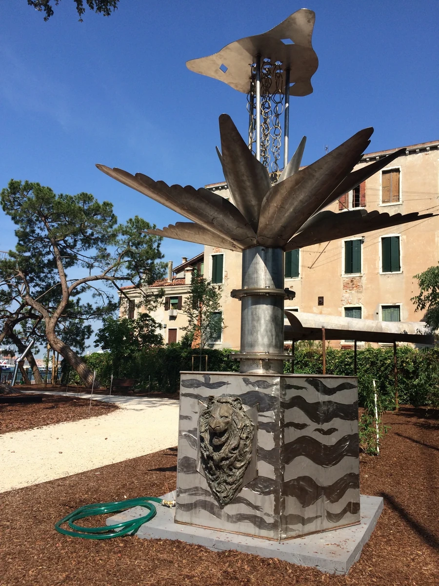 Brushed metal RainKeep sculpture with leaf canopy and hanging chain on a lion-relief base, installed in a garden beside historic buildings in Venice