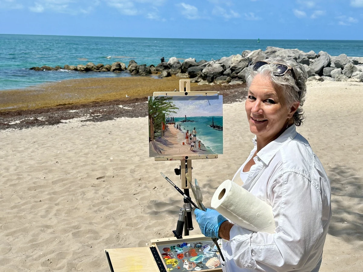 Barbara Hann painting en plein air at Fort Zachary Taylor beach, at her easel with palette in hand, turned toward the camera