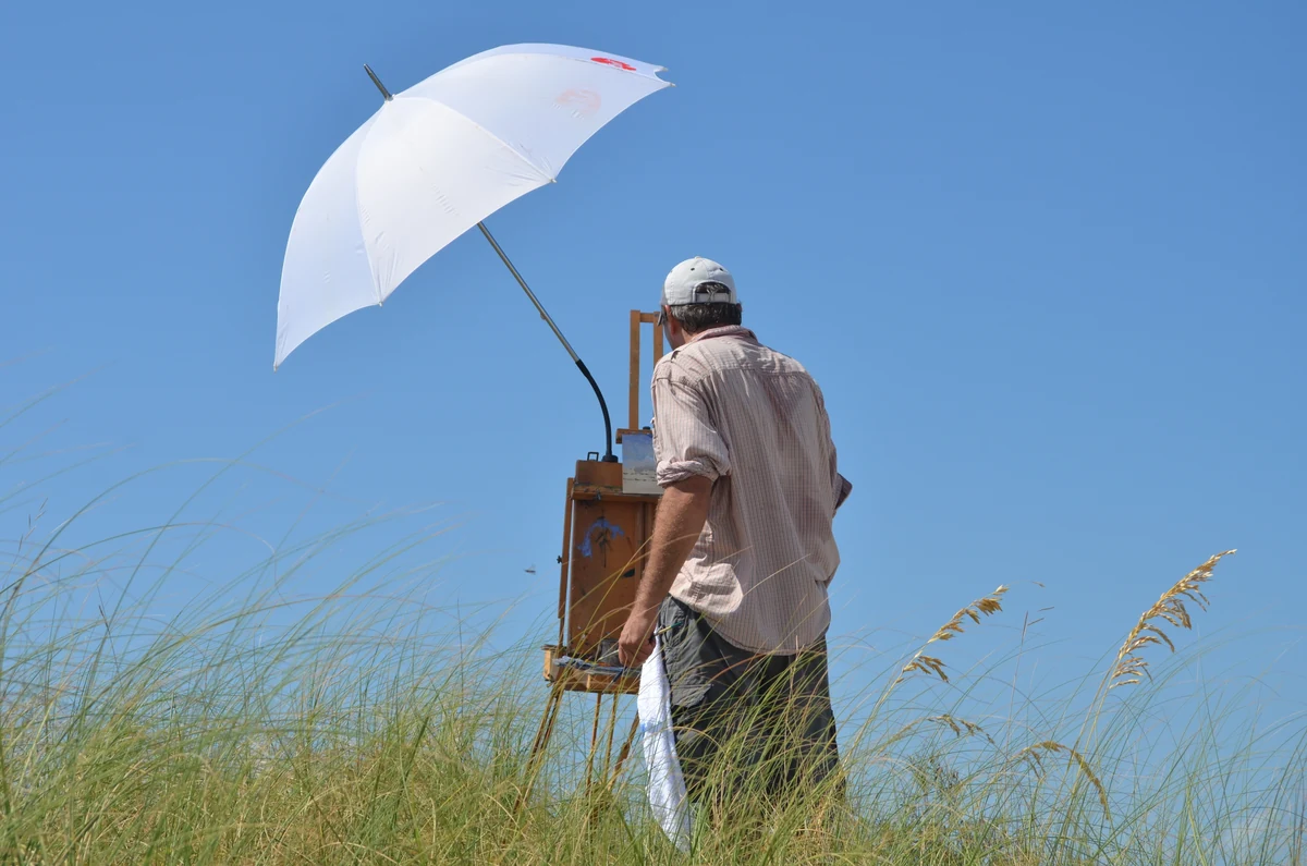 Billy Solitario painting en plein air at his easel in the dunes under a white umbrella, seen from behind