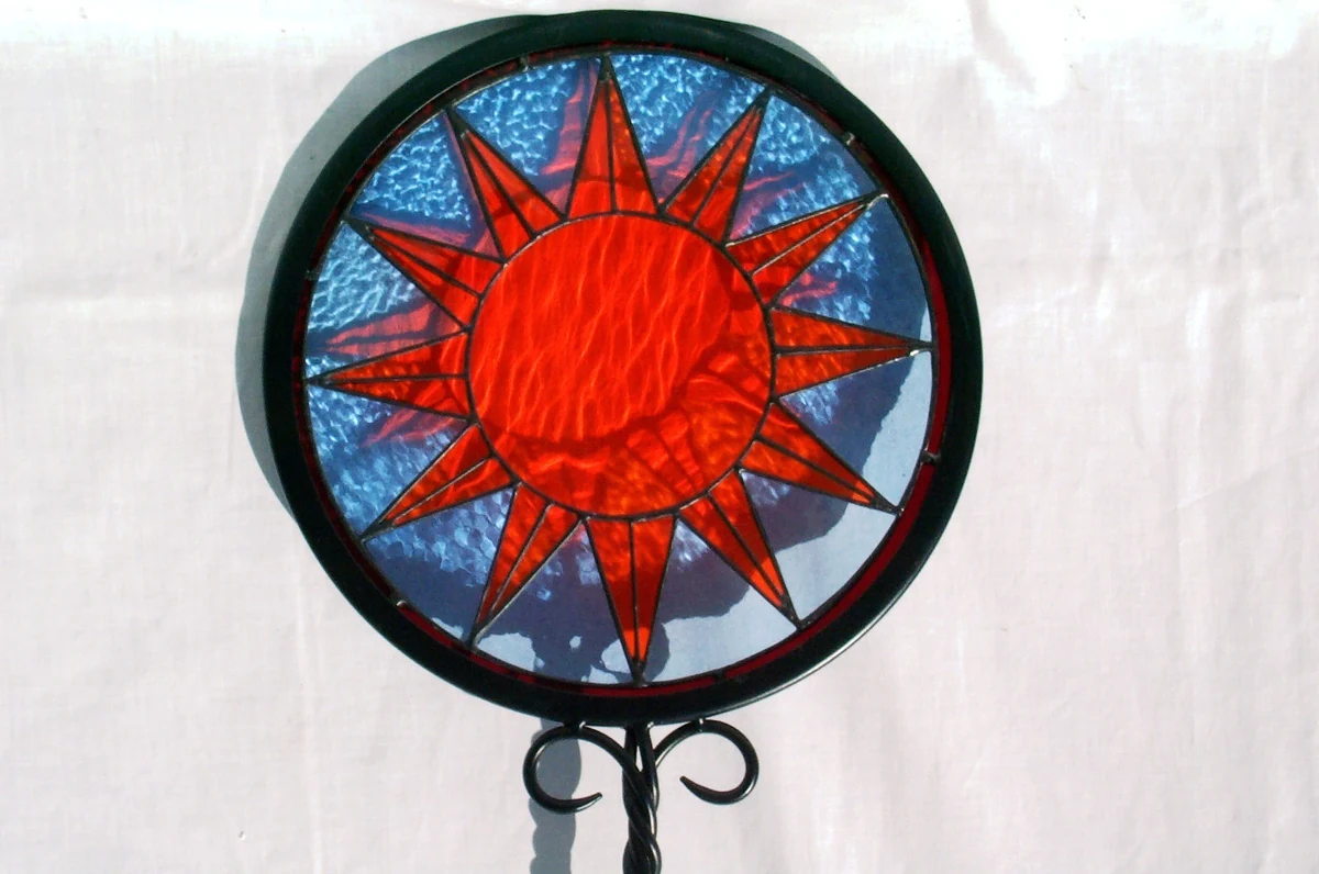 Circular stained glass garden stake panel with a red rayed sun on a mottled blue sky, set in a dark metal ring on a scrolled iron stand