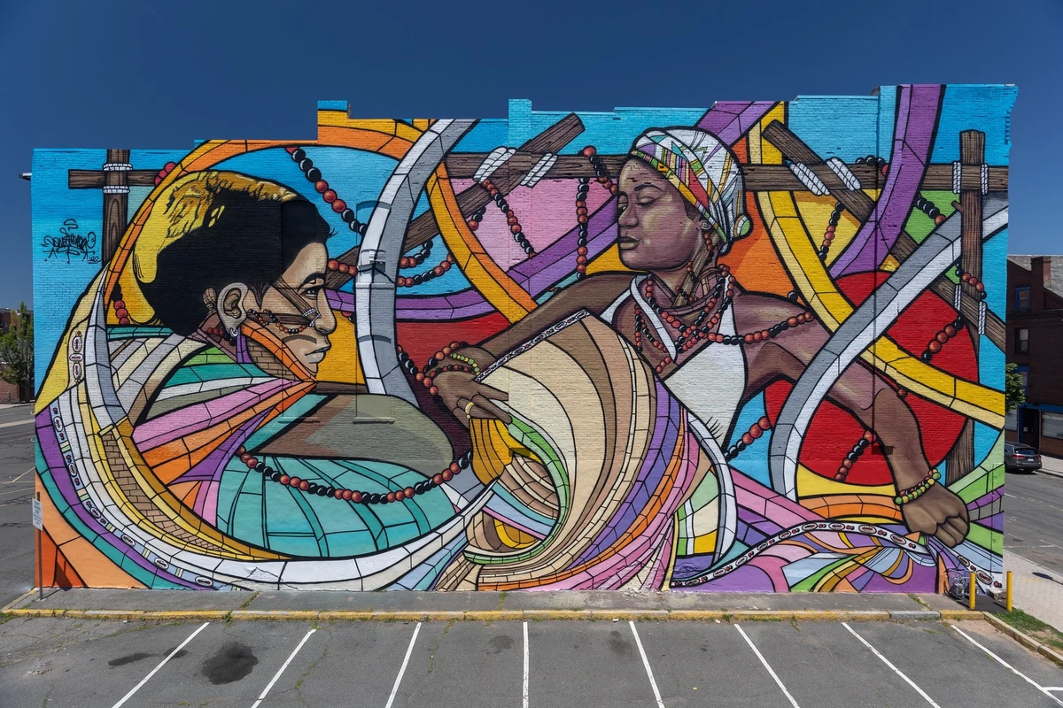 Tun Cutum PA, exterior wall mural of two figures in profile among interlaced wooden beams, red beads and bands of color, against a clear blue sky
