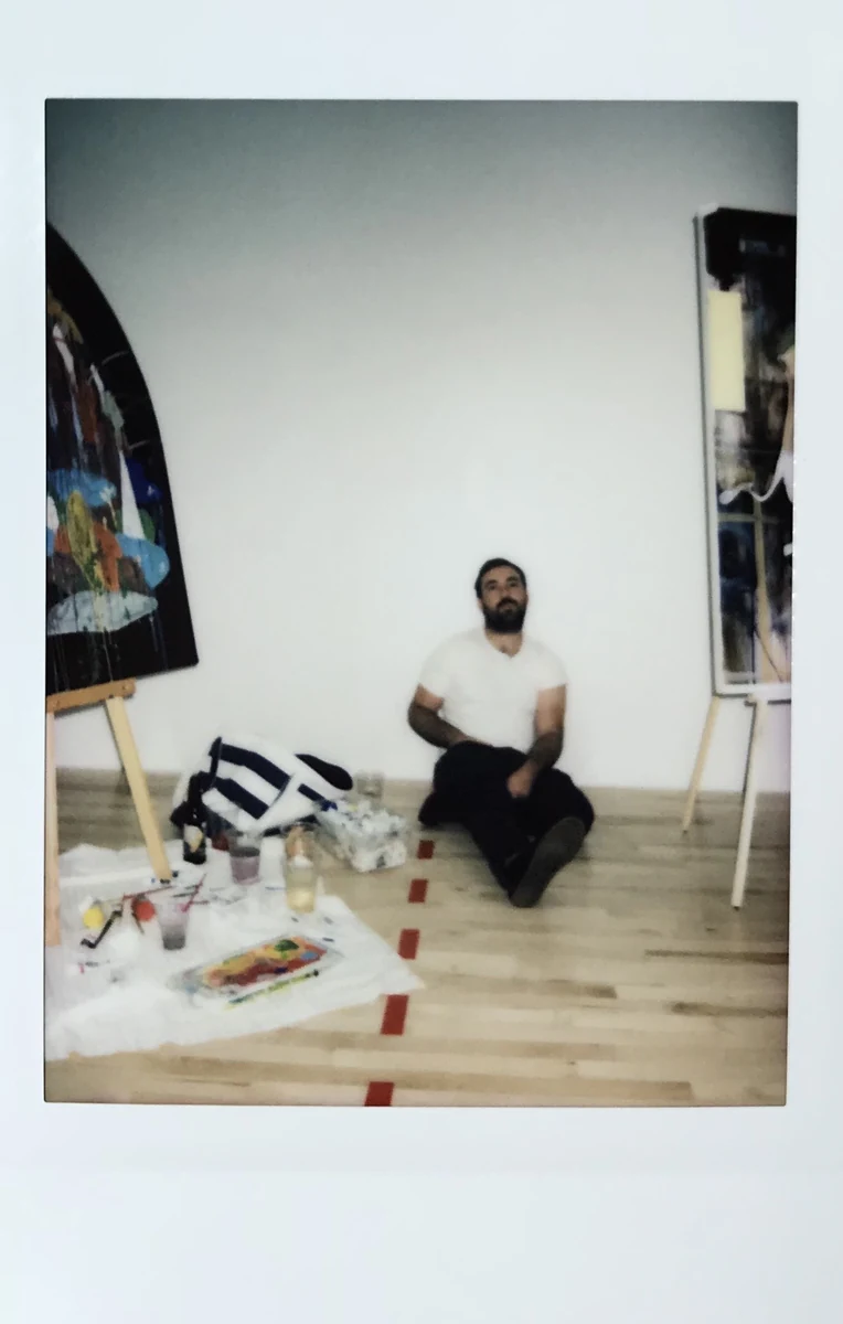 Gregory Dirr seated on his studio floor between two easels holding paintings in progress