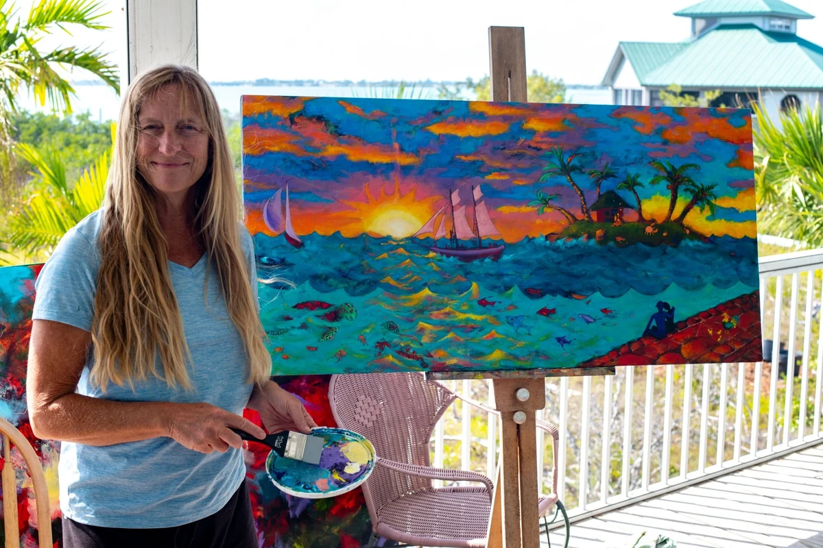 Jane Wood on a Keys porch holding a palette and brush beside her painting "Sunset Key" on an easel
