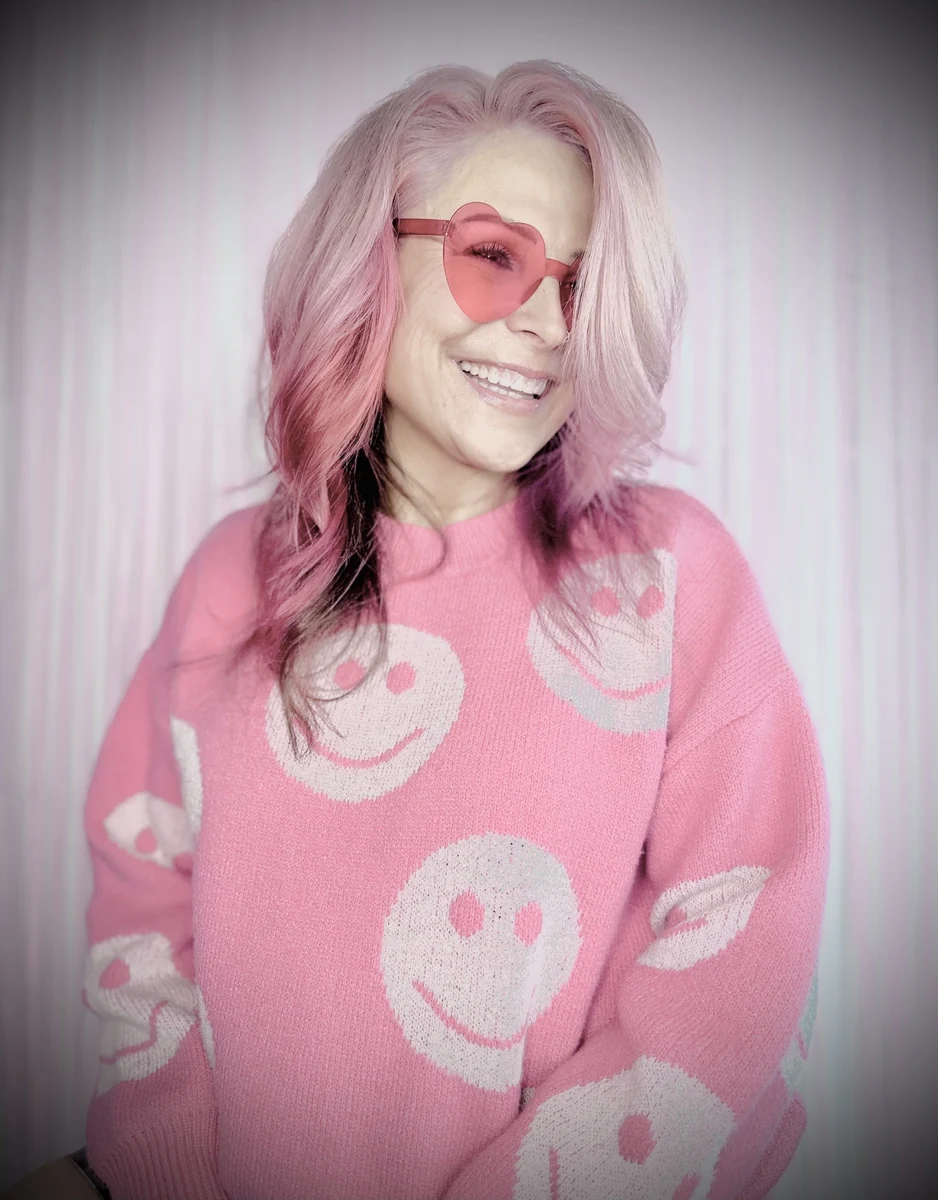 Jennifer Stephens, the bio portrait from her About page: smiling, long pink-toned hair, heart-shaped pink sunglasses, pink smiley-face sweater, against a pale curtain