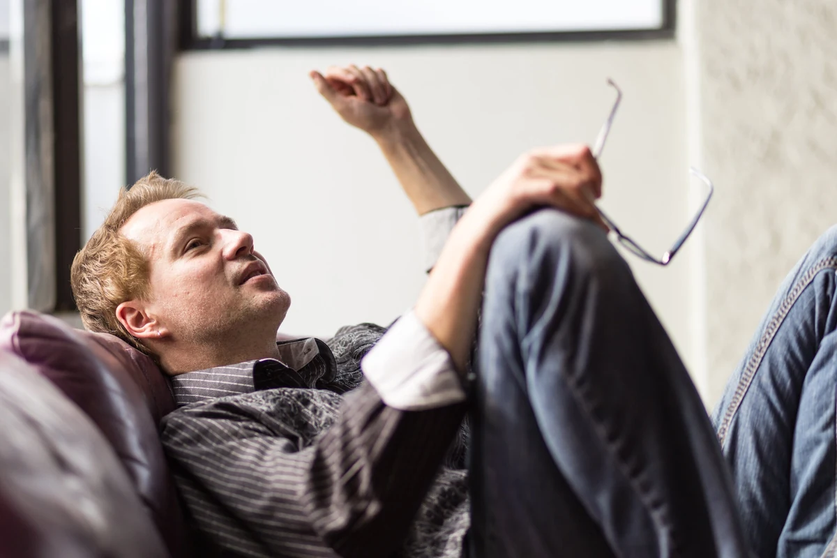 Jonathan Brown, reclining against a couch by a studio window with his glasses in hand