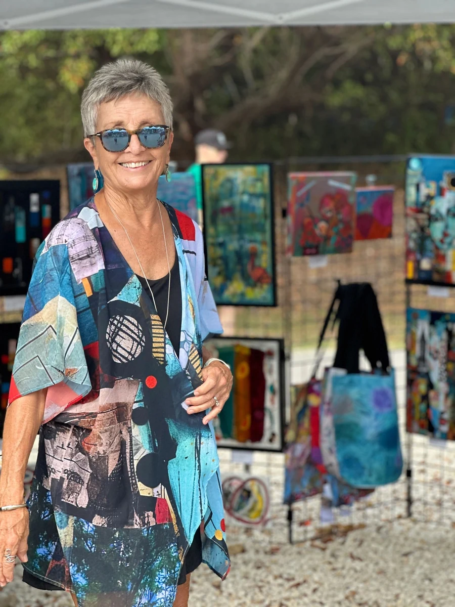 Kathy Sperl-Bell smiling in her festival booth, wearing one of her own printed ArtWear pieces, with her paintings and tote bags hung on the display panels behind her