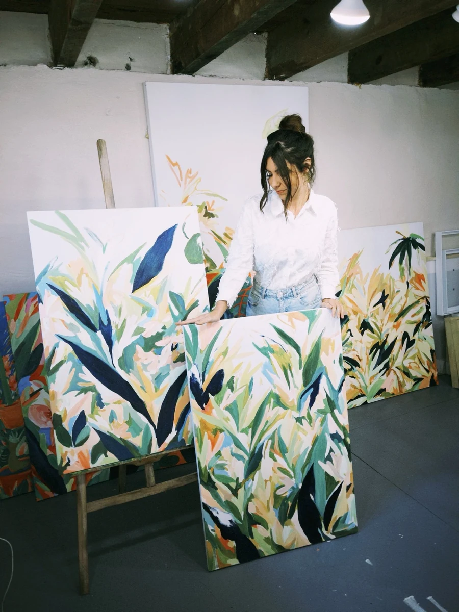 Katlin Spain in her studio, holding one of her large tropical foliage paintings with two more on the wall behind her