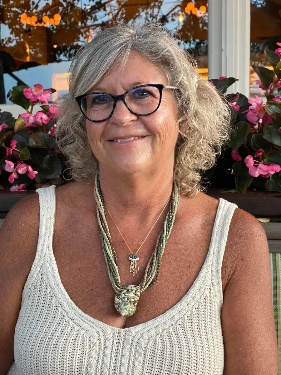Kelly Walkotten, smiling portrait outdoors at dusk beside pink begonias, wearing a shell and beaded necklace