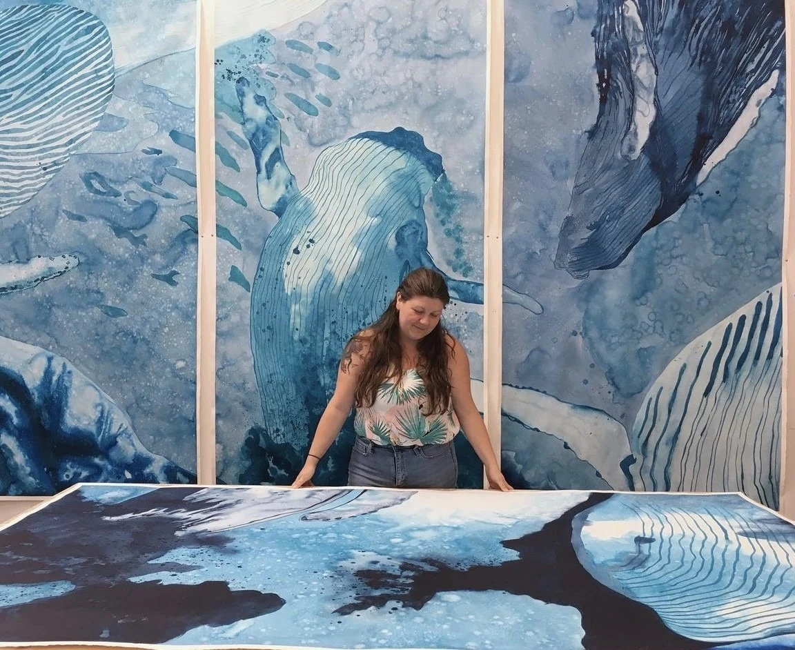 Lauren Boilini in her studio, standing at a large work on paper laid flat, in front of a four-panel blue painting