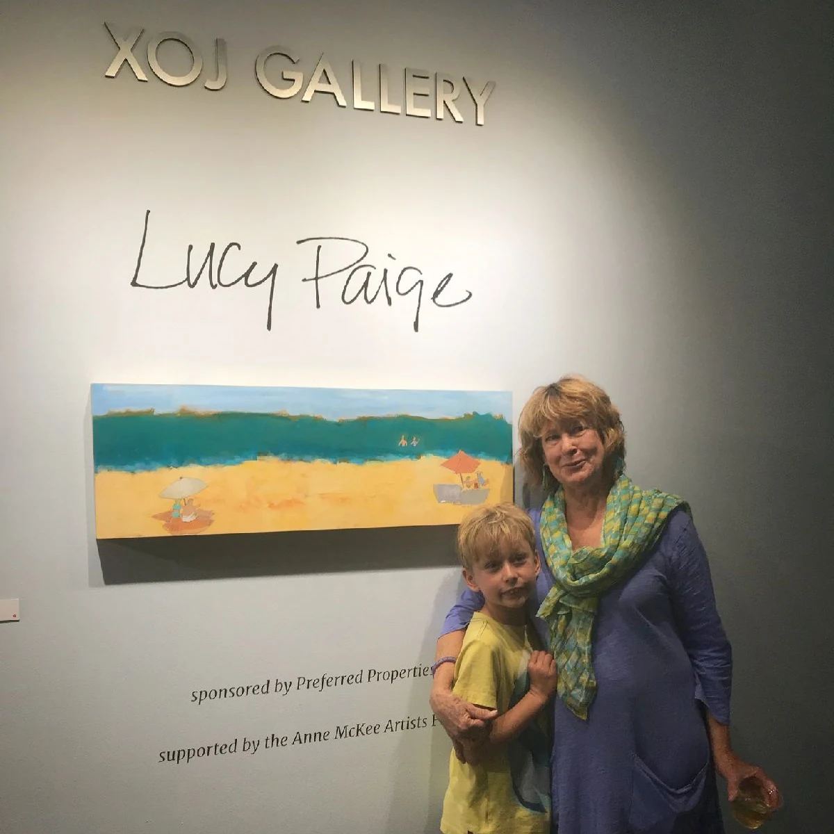 Lucy Paige at her solo exhibition, standing beside one of her beach paintings under the gallery wall lettering of her name, with a young child beside her