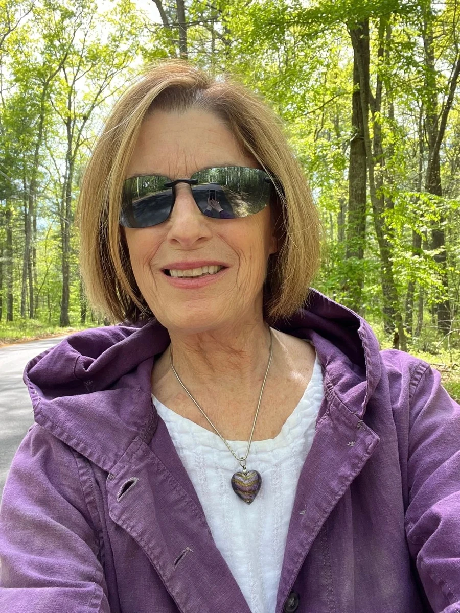 Lynne Vantriglia outdoors on a tree-lined road, in a purple jacket and sunglasses
