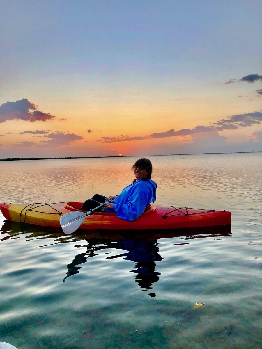 Nancy Adams seated in a red kayak on shallow Florida Keys water at sunset, turning to smile at the camera