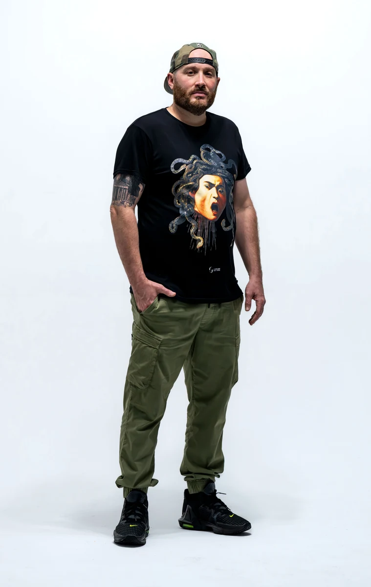 Shea Monahan, full length studio portrait against a white backdrop, wearing a t-shirt printed with his own Mask of Medusa painting