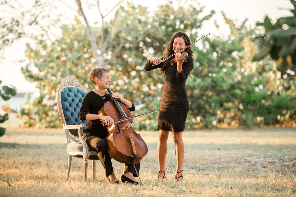 The duo performing outdoors at golden hour, Denise Nathanson playing cello seated in an upholstered chair while Irie Monte plays violin standing beside her