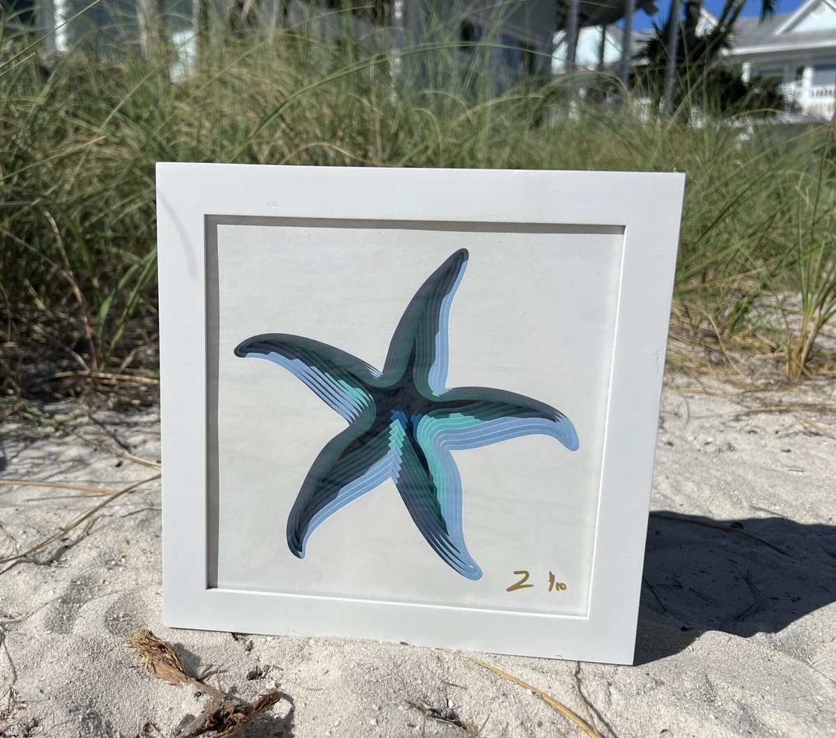 Starfish, a layered laser-cut starfish in blue and teal in a white shadow-box frame, photographed on the sand