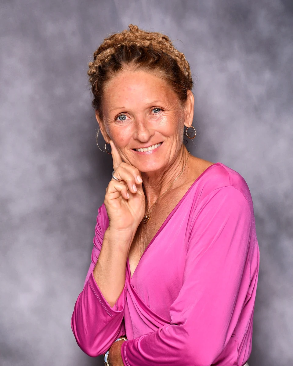 Studio portrait of Vicki Grace Boguszewski, smiling, hand to her cheek, in a magenta top against a gray backdrop